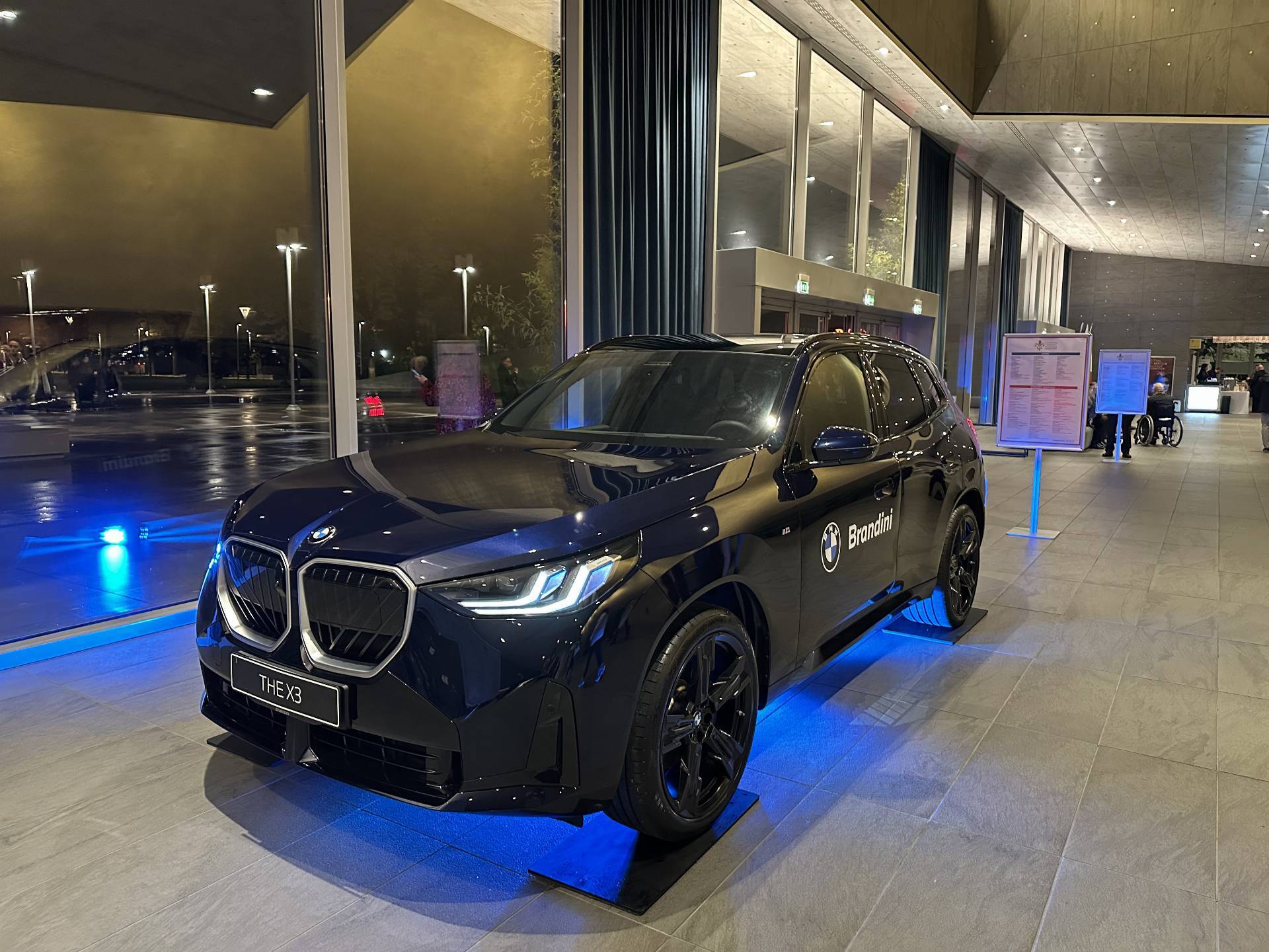 BMW X3 esposta presso il Teatro del Maggio Musicale Fiorentino, con logo Brandini, illuminata da luci blu in un ambiente moderno ed elegante.